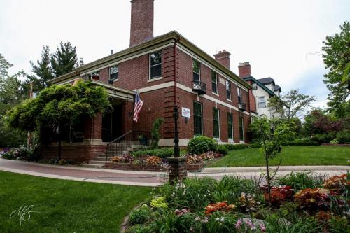 A brick house with a porch, surrounded by lush gardens and colorful flowers, under a cloudy sky. An American flag is visible.