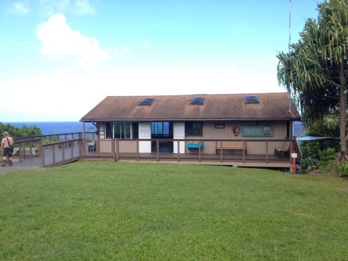 A wooden visitor center with a sloped roof, surrounded by green grass and overlooking the ocean under a blue sky.
