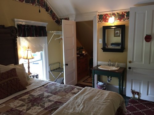 Cozy bedroom with a quilted bed, vintage decor, a lamp, and a small sink area, featuring warm yellow walls.