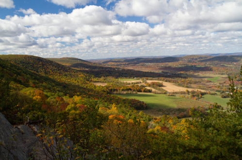 A scenic view of rolling hills and valleys, adorned with autumn foliage under a partly cloudy sky.