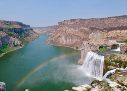 A scenic view of a waterfall cascading into a river, surrounded by cliffs and a rainbow in the mist.