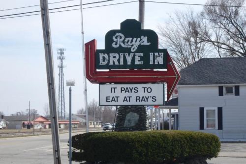 Sign for Ray's Drive-In with an arrow pointing left, stating "It pays to eat at Ray's." Surrounding landscape visible.