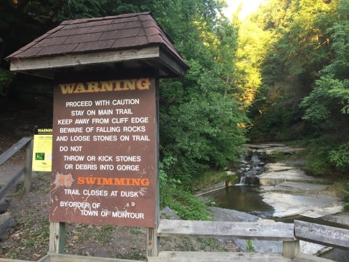 A warning sign at a nature trail advises caution, swimming restrictions, and safety tips near a wooded area and stream.