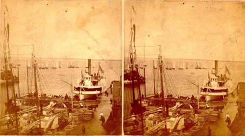 Historic sepia-toned image of a busy harbor with a steamboat and sailboats in the background.