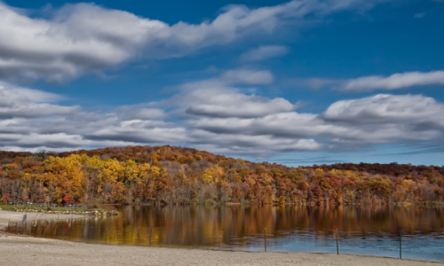 A serene lake surrounded by autumn foliage and a cloudy blue sky, reflecting vibrant fall colors on the water.