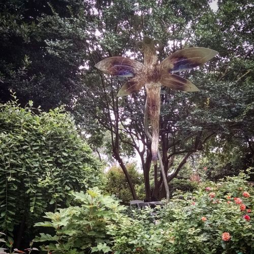 A large, translucent butterfly sculpture stands among lush greenery and blooming flowers in a garden setting.