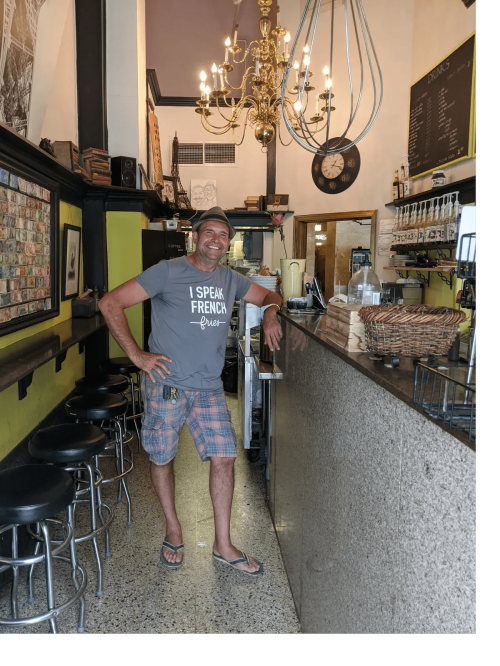 A smiling man in a casual t-shirt stands by a bar in a cozy café with a chandelier and stools.