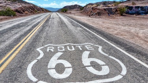 A view of a deserted road with a painted Route 66 sign on the asphalt, surrounded by arid landscape and mountains.