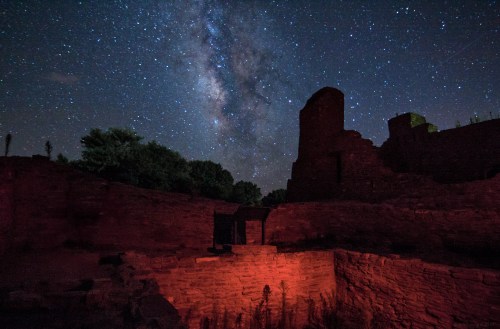 A starry night sky with the Milky Way over ancient stone ruins, illuminated by a soft red light.