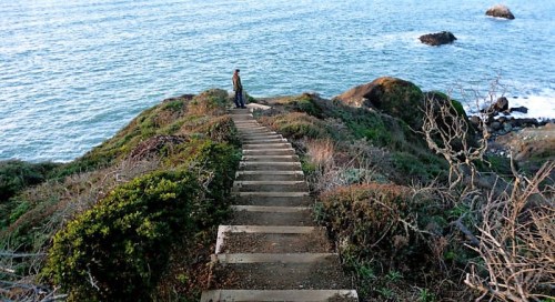 A person stands at the top of a staircase leading down to a rocky coastline with waves crashing against the shore.