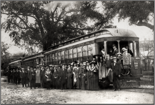 A historic black-and-white photo of a large group of people gathered by a vintage streetcar.