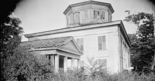 An old, weathered house with a distinctive octagonal tower, surrounded by overgrown vegetation.