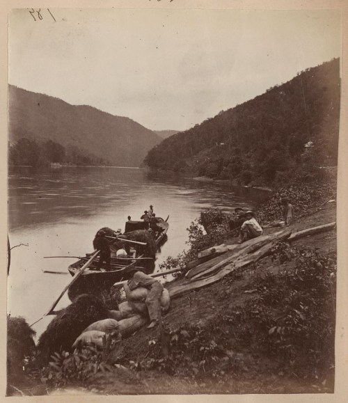 A historic black-and-white photo of a river scene with boats and people along the shore, surrounded by hills.