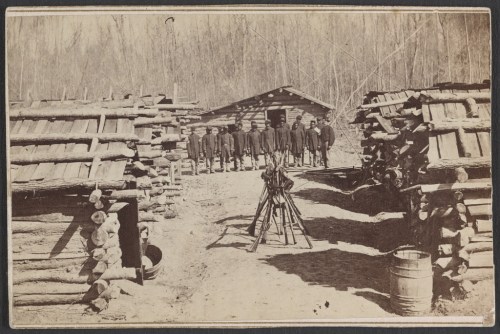 A historic photo of soldiers in uniform standing in front of log cabins, with a camera on a tripod in the foreground.