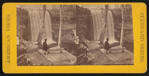 A historic photo of a person sitting on a bench near a waterfall, surrounded by trees and rocky terrain.