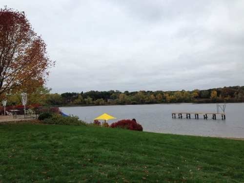 A serene lakeside view with autumn foliage, a yellow tent, and a wooden dock under a cloudy sky.