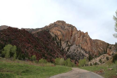 A winding road leads through a lush green valley, flanked by rugged, colorful mountain cliffs under a cloudy sky.