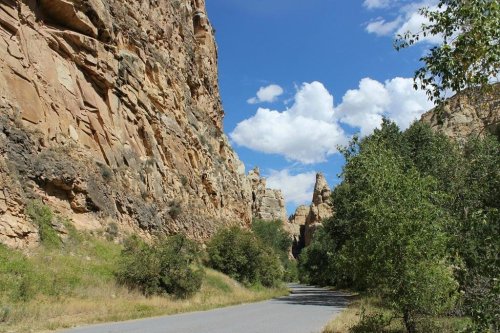 A scenic view of a winding road surrounded by tall rock formations and greenery under a blue sky with fluffy clouds.