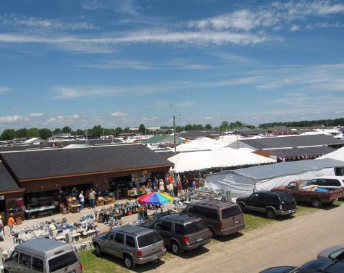 A bustling outdoor market with tents, vendors, and parked vehicles under a blue sky with scattered clouds.