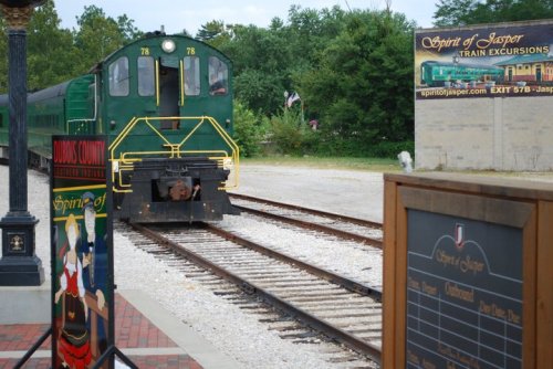 A green train approaches a station with signs for Spirit of Jasper Train Excursions and nearby trees in the background.