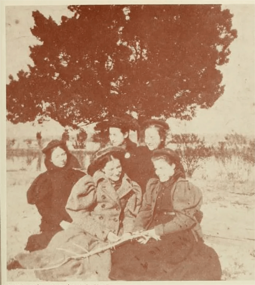 Five women in vintage clothing pose together outdoors under a large tree, smiling and enjoying each other's company.