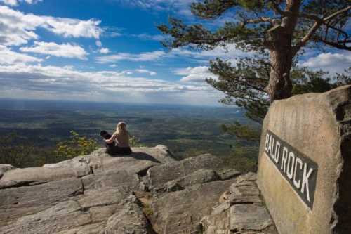 A person sits on a rocky ledge at Bald Rock, overlooking a vast landscape under a blue sky with clouds.