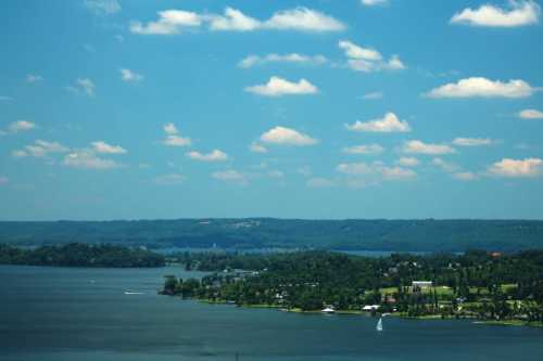 A serene lake view with lush green shores, dotted with trees, under a bright blue sky with fluffy white clouds.