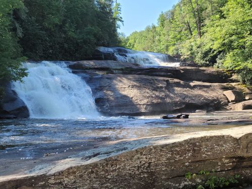 A serene waterfall cascades over rocky ledges, surrounded by lush green trees under a clear blue sky.