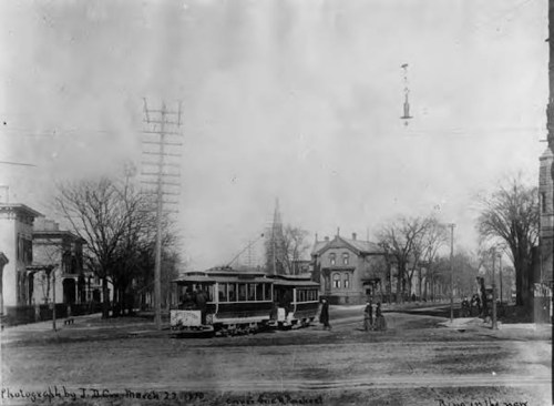 Historic black-and-white photo of a streetcar at an intersection, with trees and buildings in the background.