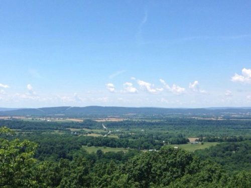 A panoramic view of lush green valleys and distant mountains under a clear blue sky with scattered clouds.