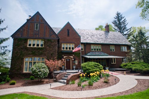 A large brick house with a slate roof, surrounded by greenery and flowers, featuring an American flag on the porch.