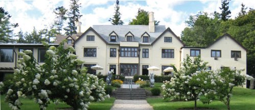 A large, elegant house surrounded by blooming white flowers and lush greenery under a blue sky.