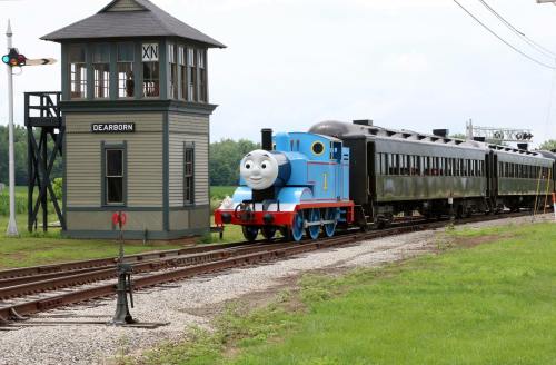 A blue train resembling Thomas the Tank Engine approaches a vintage station with a green landscape in the background.