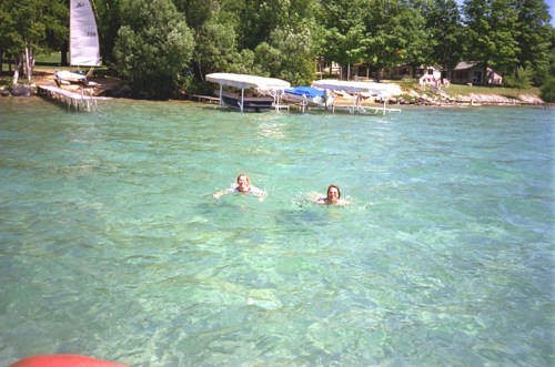 Two people swim in clear, turquoise water near a dock with boats and trees in the background.