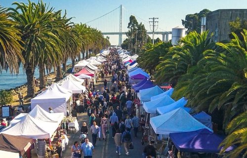 A bustling outdoor market lined with tents, palm trees, and a bridge in the background, filled with people exploring.