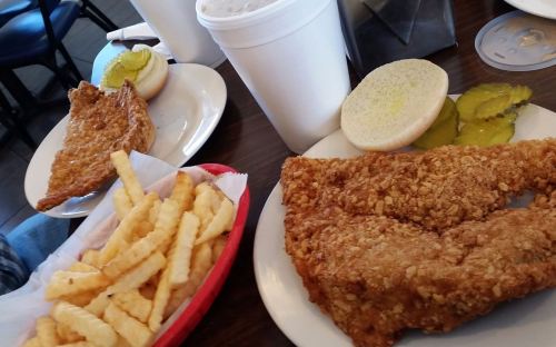 A table with two plates of fried fish, pickles, a bun, and a side of crinkle-cut fries, with a drink in a cup.