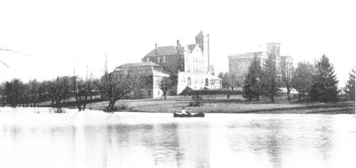 Historic black-and-white image of a large building by a river, with trees and a small boat in the foreground.