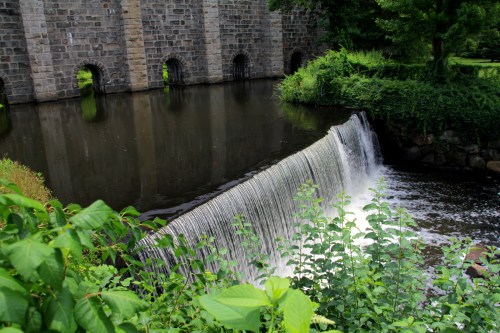 A serene waterfall cascades over a stone dam, surrounded by lush greenery and reflecting in the calm water below.