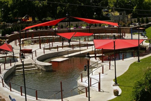 A landscaped area with a winding water feature, shaded by red canopies, surrounded by green grass and flowers.