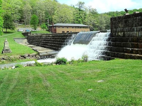 A waterfall cascades over stone steps beside a grassy area and a building surrounded by trees.
