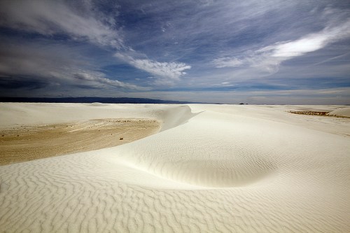 A vast expanse of white sand dunes under a cloudy sky, showcasing rippled textures and gentle slopes.