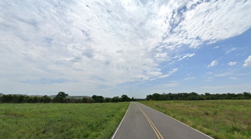 A wide, empty road stretches through a green landscape under a partly cloudy sky.