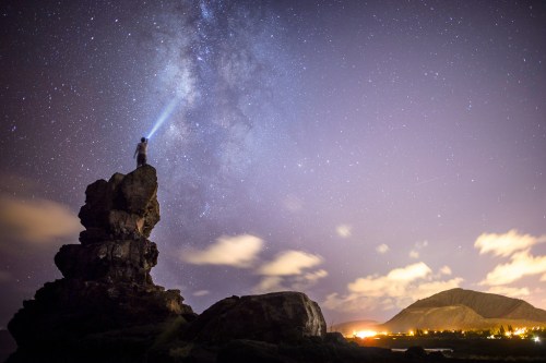 A person stands on a rock, shining a flashlight at the starry night sky, with mountains and lights in the background.