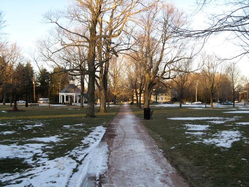 A peaceful park path lined with bare trees, leading to a gazebo, with patches of snow on the ground.