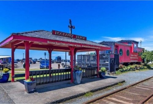 A colorful gazebo at Rockaway Beach, with a sign and decorative planters, overlooking a parking area and ocean.