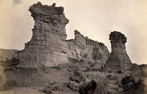 Black and white photo of towering rock formations in a rugged landscape, with a person standing near the base.