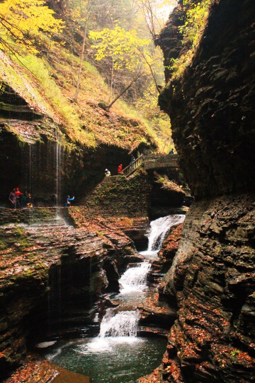 A scenic gorge with waterfalls, surrounded by rocky cliffs and autumn foliage, with people exploring the area.