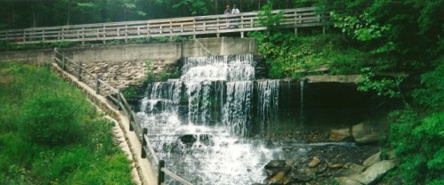 A serene waterfall cascades over rocks, with a wooden bridge and lush greenery in the background.