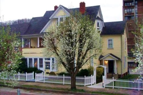 A charming yellow house with white trim, surrounded by blooming trees and a white picket fence.