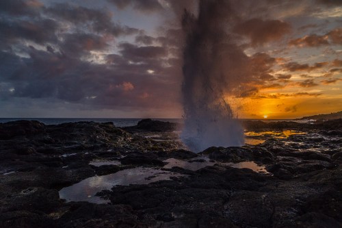 A dramatic sunset over rocky shores, with water spouting up from the rocks against a colorful sky.